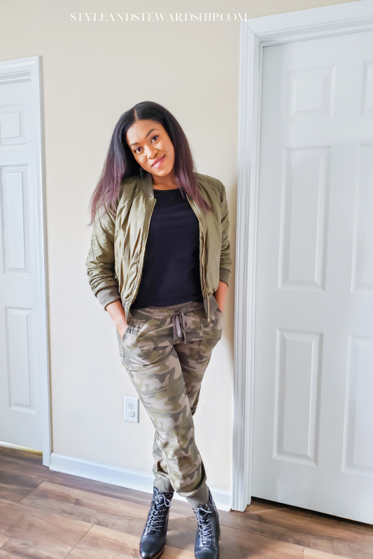 Woman in a black top, green jacket with hands in pockets of camouflage pants.