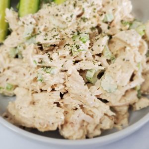 Large white bowl with chunky chicken salad and cucumber pickle spears next to chicken salad style and stewardship dot com