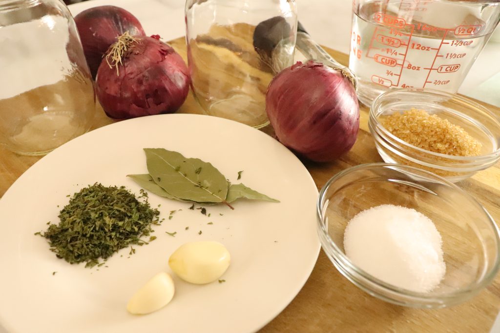 dried seasonings on a white plate along with 2 garlic cloves and 3 red onions, and glass jars next to plate and small glass bowls with sugar and salt