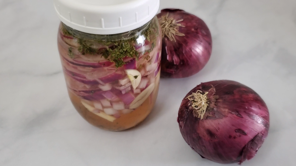 glass jar with liquid and slices of red onions layered with herbs on a counter with two whole red onions beside it