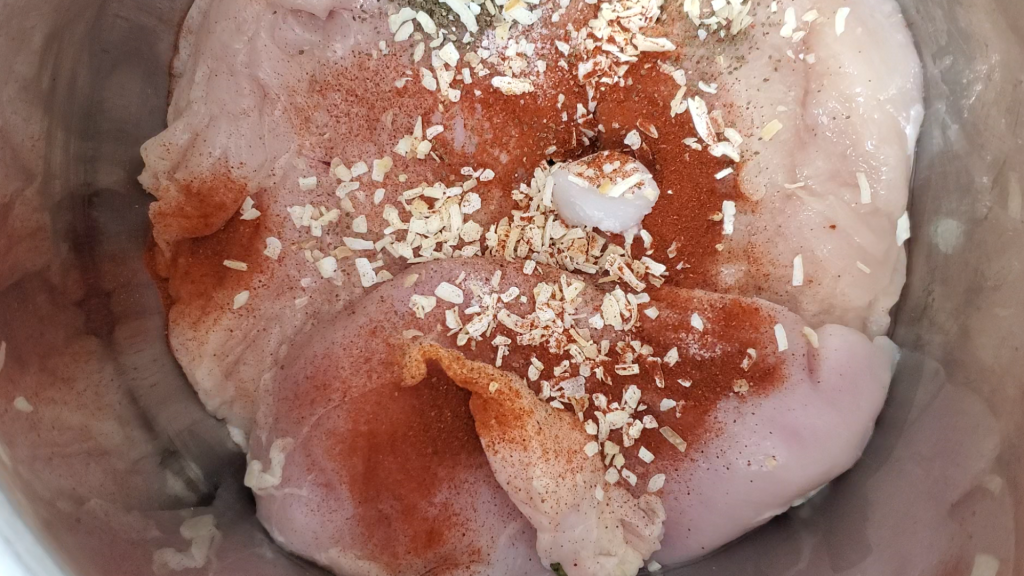 raw chicken breast in the bottom of a stainless instant pot covered with dried seasoning and coconut oil