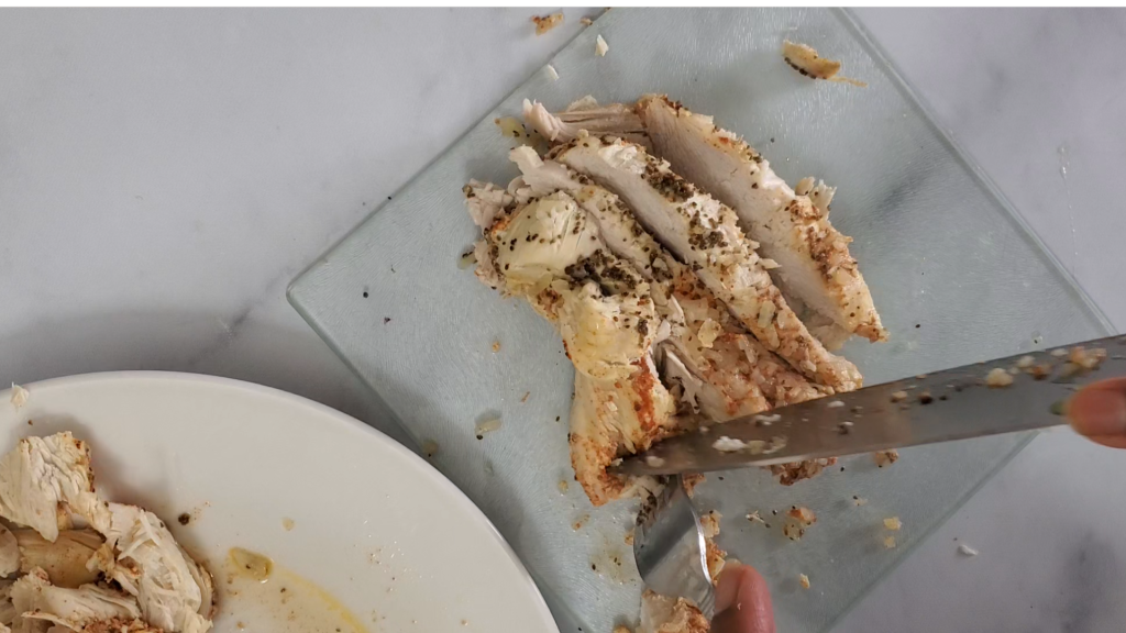 a glass cutting board with a cooked chicken breast coated in seasoning with a knife and fork being cut into chunks