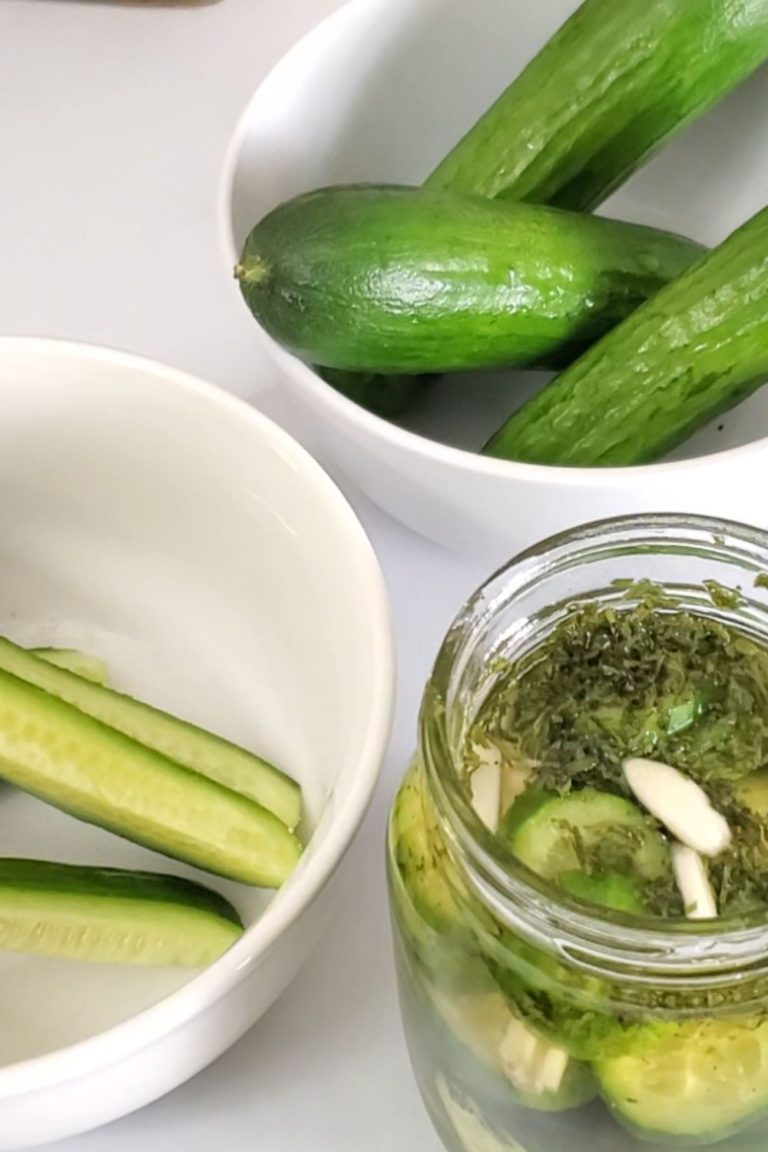 glass jar with slices of pickles, liquid and seasonings surrounded by white bowl with cucumber spears and a white bowl with whole small cucumbers