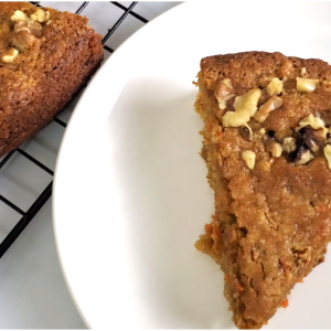 a slice of carrot cake with topped with walnuts on a small white plate. Cooling rack with carrot cake in the background style and stewardship dot com