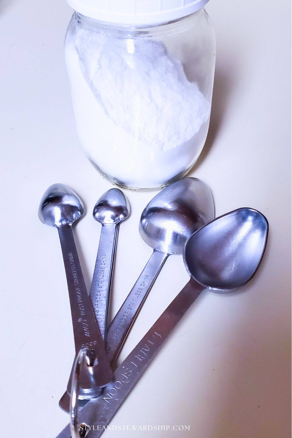 glass jar with white baking powder and silver measuring spoons fanned out in front style and stewardship dot com