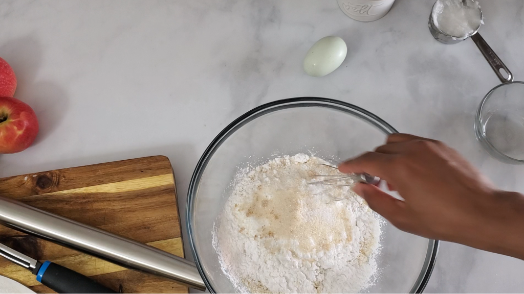 Picture of apples, a rolling pin, measuring cups, spoons, cutting board with a glass bowl filled with flour mixture and a hand mixing with a whisk