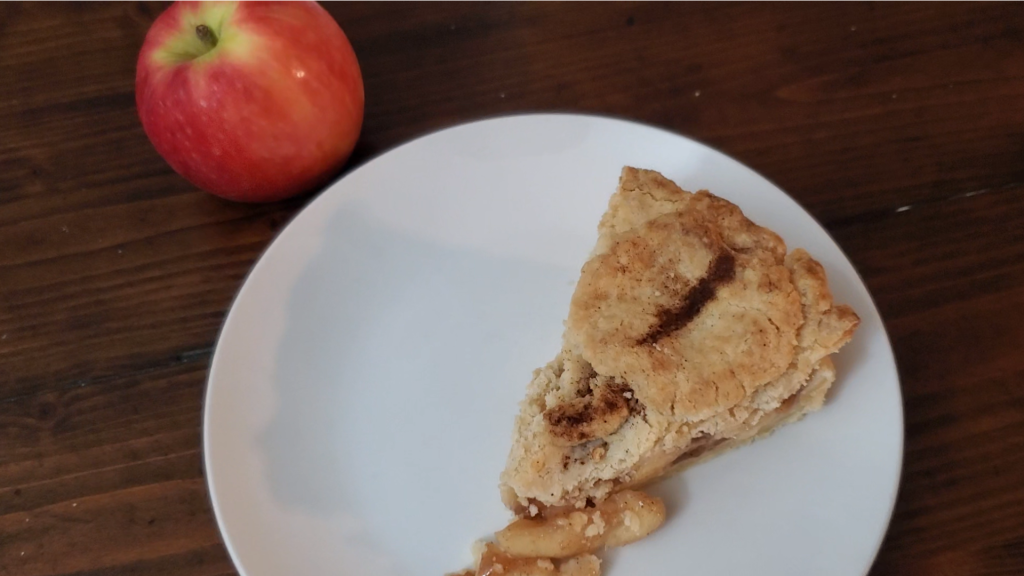 Apple pie slice on a white plate pictured with a red apple in the background