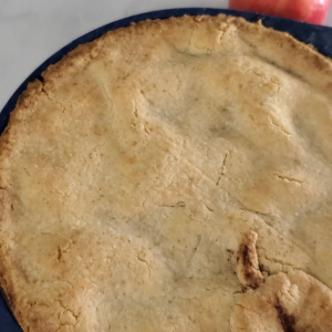 Golden Apple pie in a pie pan with blue rim and fresh apple in background