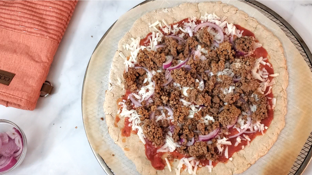 Pizza on a marble counter with ground beef, red onions and cheese on top of parchment. Sliced onions and an oven mitten are next to it. 