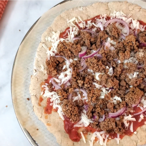 Pizza on a marble counter with ground beef, red onions and cheese on top of parchment. Sliced onions and an oven mitten are next to it.