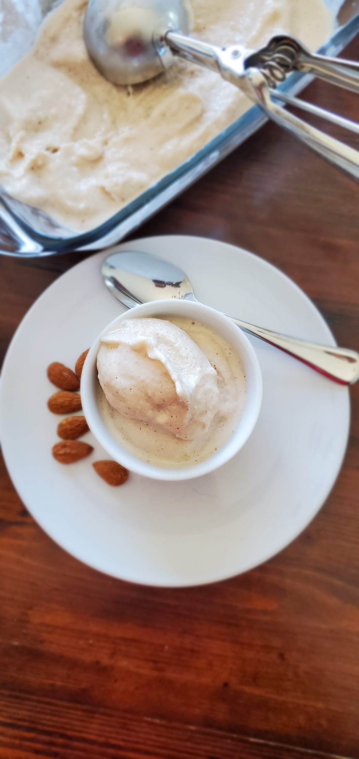 Almond milk ice cream in a small white bowl on top of a small white plate surrounded by whole almonds