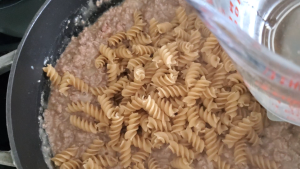 measuring glass with water being added to a cast iron pan of sauce, dry noodles, and beef