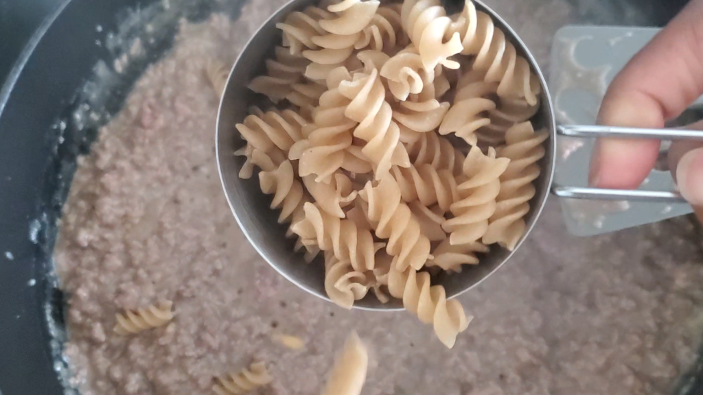 one cup of dry rotini noodles being added to a meat a sauce mixture
