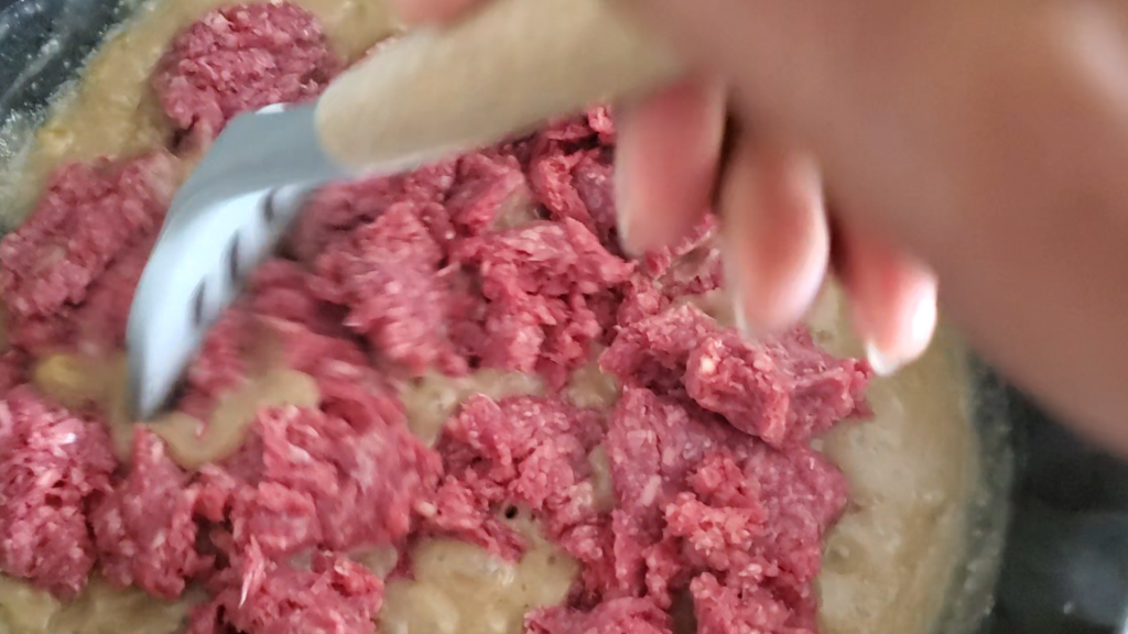 raw ground beef being added to a pan with sauce and a hand using a spatula to break the meat apart