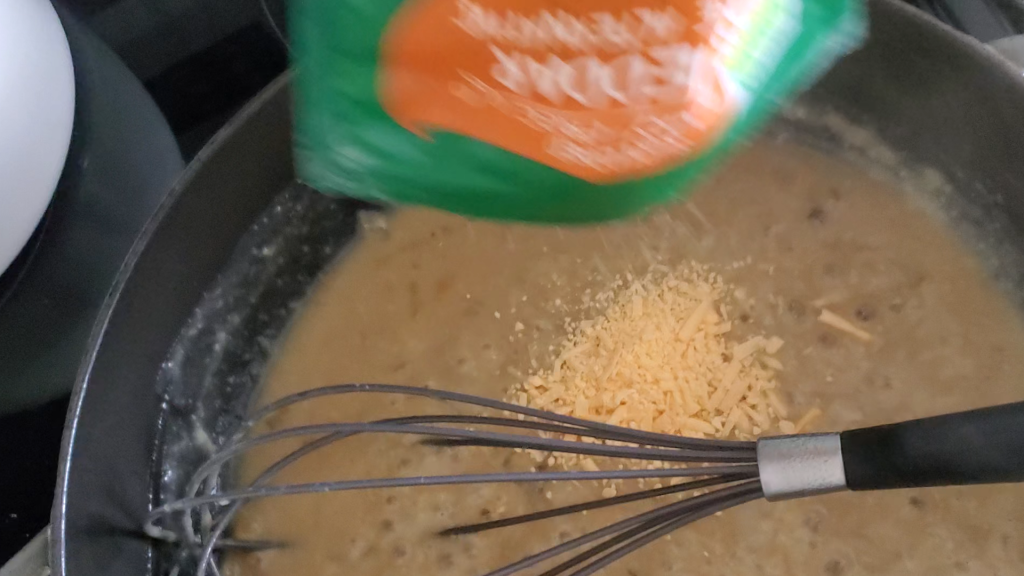 vegan cheese being added to a pan with gravy a whisk is pictured as well