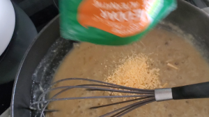 vegan cheese being added to a pan with gravy a whisk is pictured as well