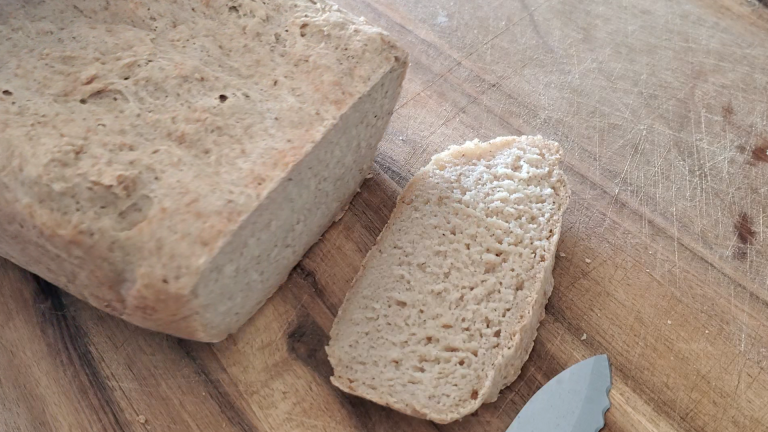 Loaf of bread on top of a large wooden cutting board and one slice is laying next to it.