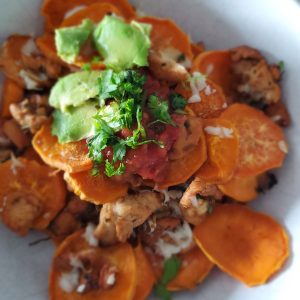 white bowl on top of a wooden cutting board, filled with sweet potato round slices, chicken, white cheese, avocado, and cilantro