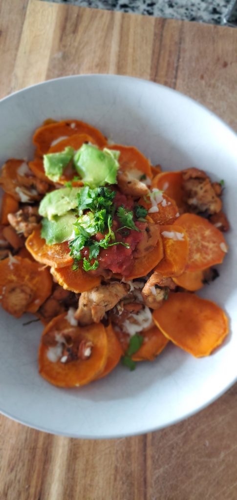 white bowl on top of a wooden cutting board, filled with sweet potato round slices, chicken, white cheese, avocado, and cilantro