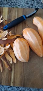 three sweet potatoes on a wooden cutting board that have been peeled. A large pile of peels lays on the board along with a black vegetable peeler