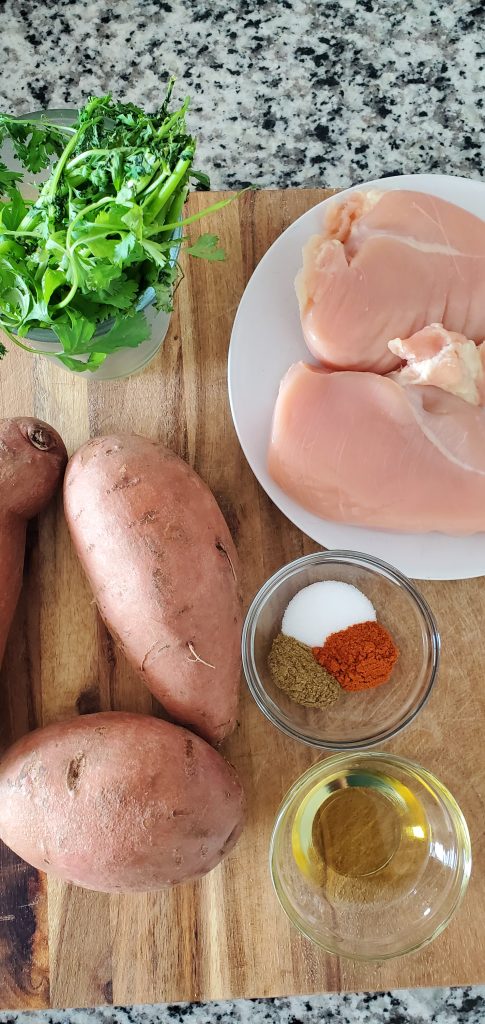 a small glass bowl with ground seasonings, 3 sweet potatoes next to it along with fresh cilantro and raw boneless skinless chicken breasts on a white plate