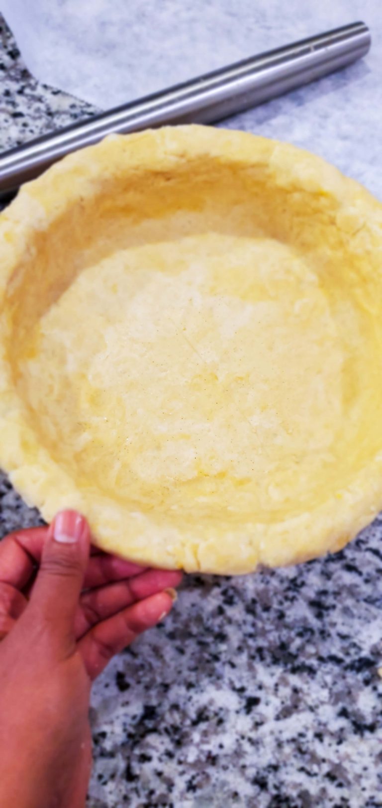 A hand is holding a pie pan and dough next to a metal rolling pin on a granite countertop