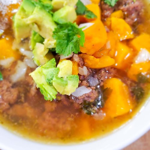 White bowl with beef, butternut squash, and broth topped with cilantro, avocado, and onion.