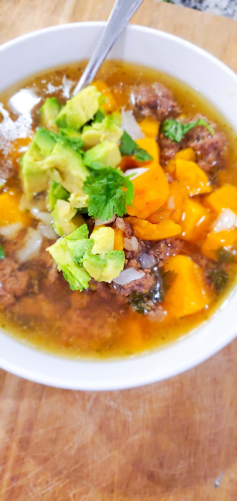 White bowl with beef, butternut squash, and broth topped with cilantro, avocado, and onion.