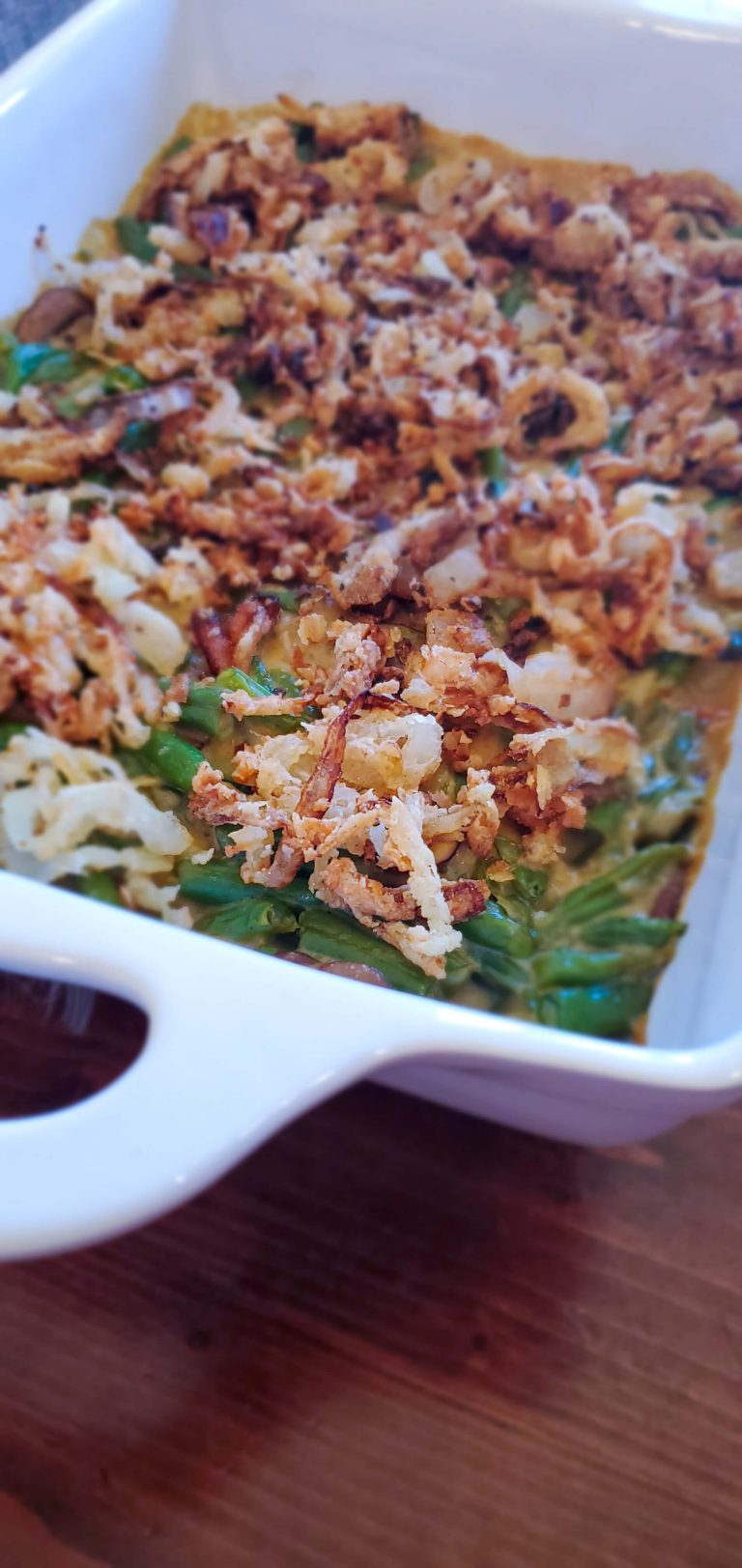 Bright green beans in a casserole dish with creamy brown sauce, topped with golden fried onions in a large white rectangular dish.