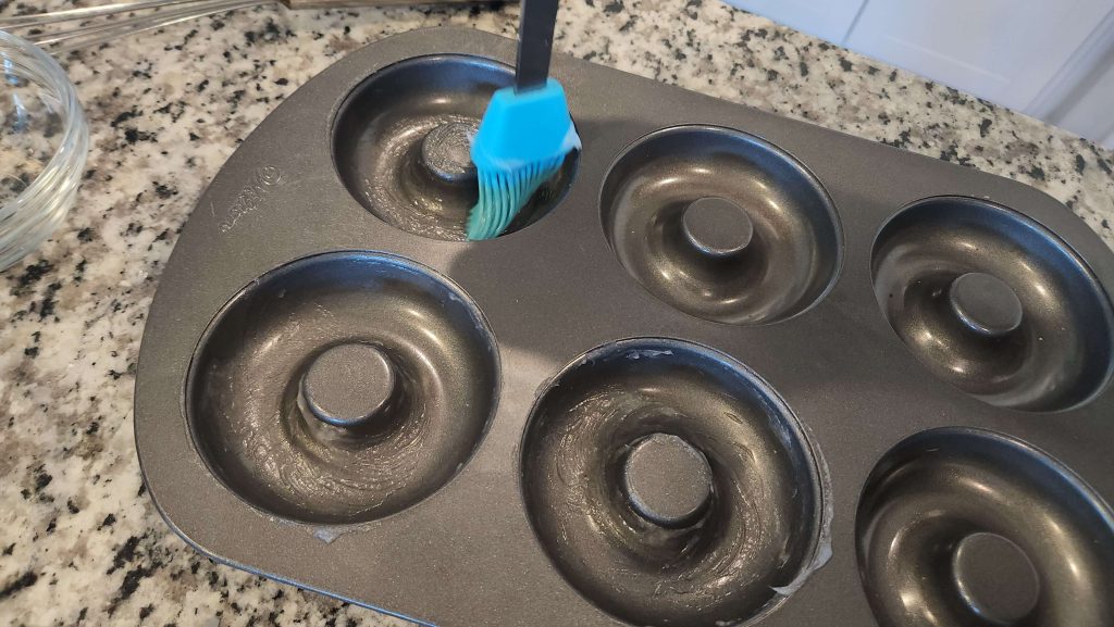 A blue silicone brush spreading coconut oil onto an empty doughnut baking pan.