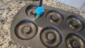 A blue silicone brush spreading coconut oil onto an empty doughnut baking pan.
