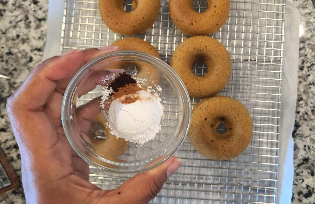 Hand holding a small glass bowl with powdered sugar and cinnamon above baked pumpkin muffins cooling on a rack below.