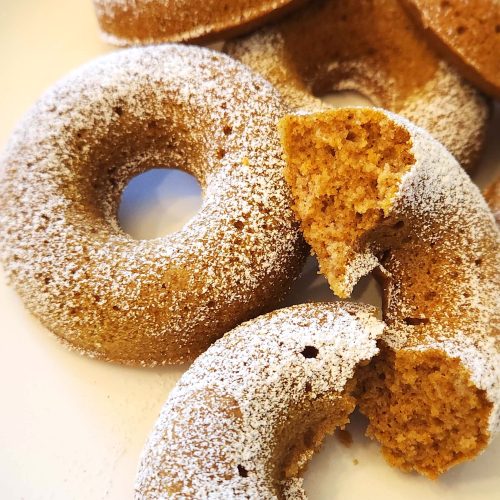 A white plate with 6 baked pumpkin doughnuts with a dusting of powdered sugar, pumpkin puree in the background