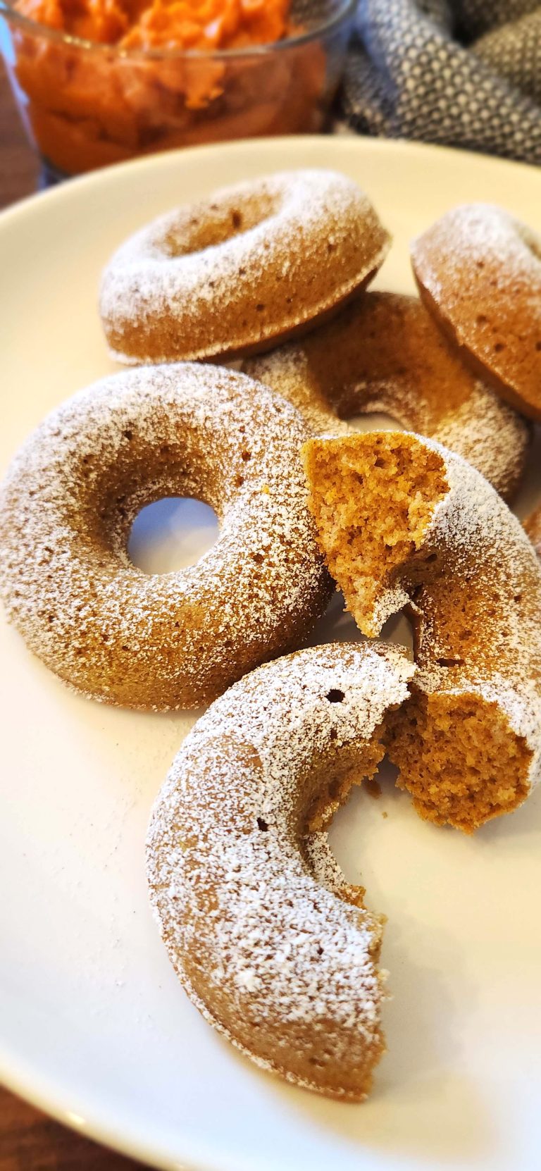 A white plate with 6 baked pumpkin doughnuts with a dusting of powdered sugar, pumpkin puree in the background