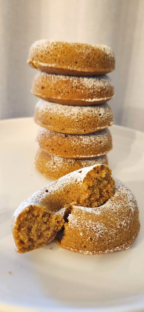 A stack of six baked gluten free doughnuts on a white plate and one split in half to show the texture.