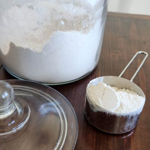 A large glass container with flour and lid sitting next to it and a silver measuring cup with flour