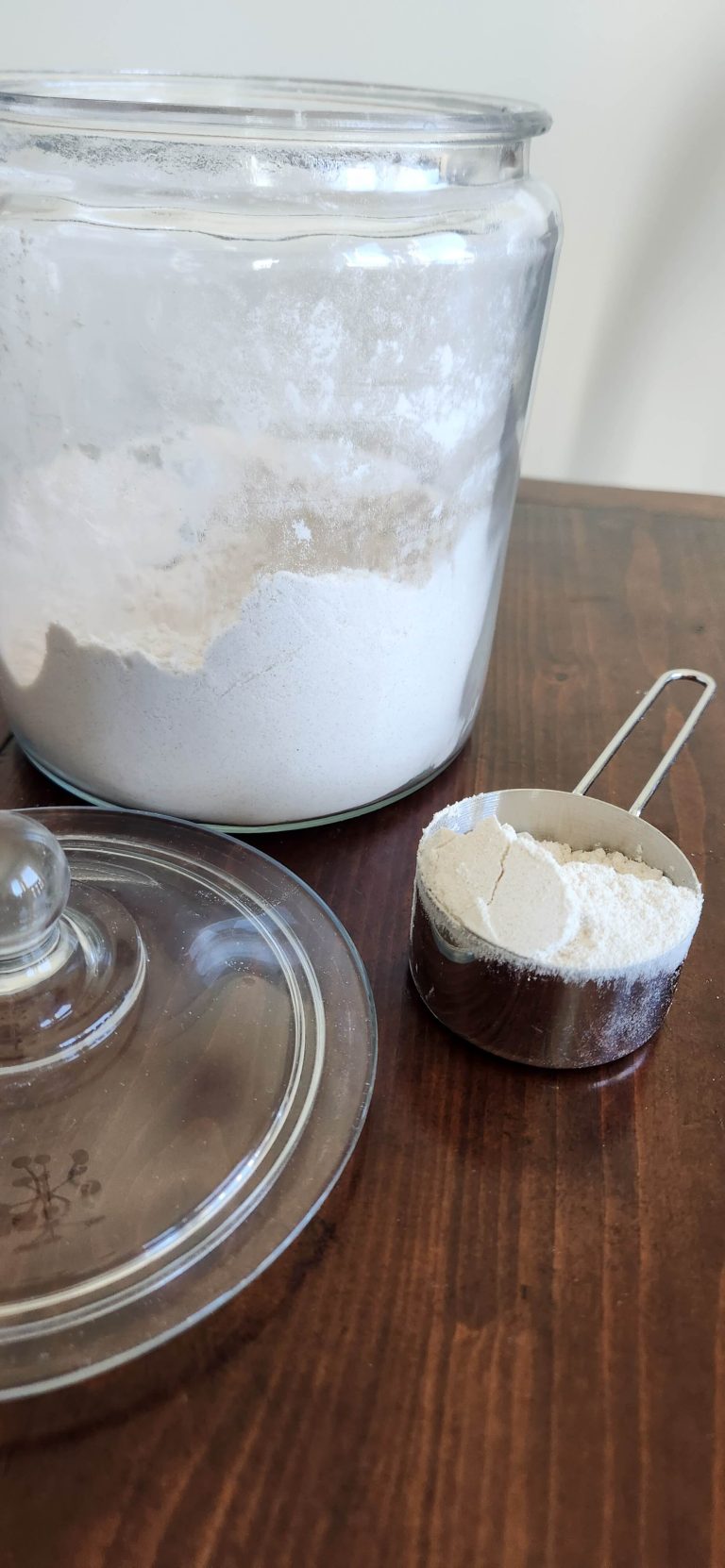 A large glass container with flour and lid sitting next to it and a silver measuring cup with flour
